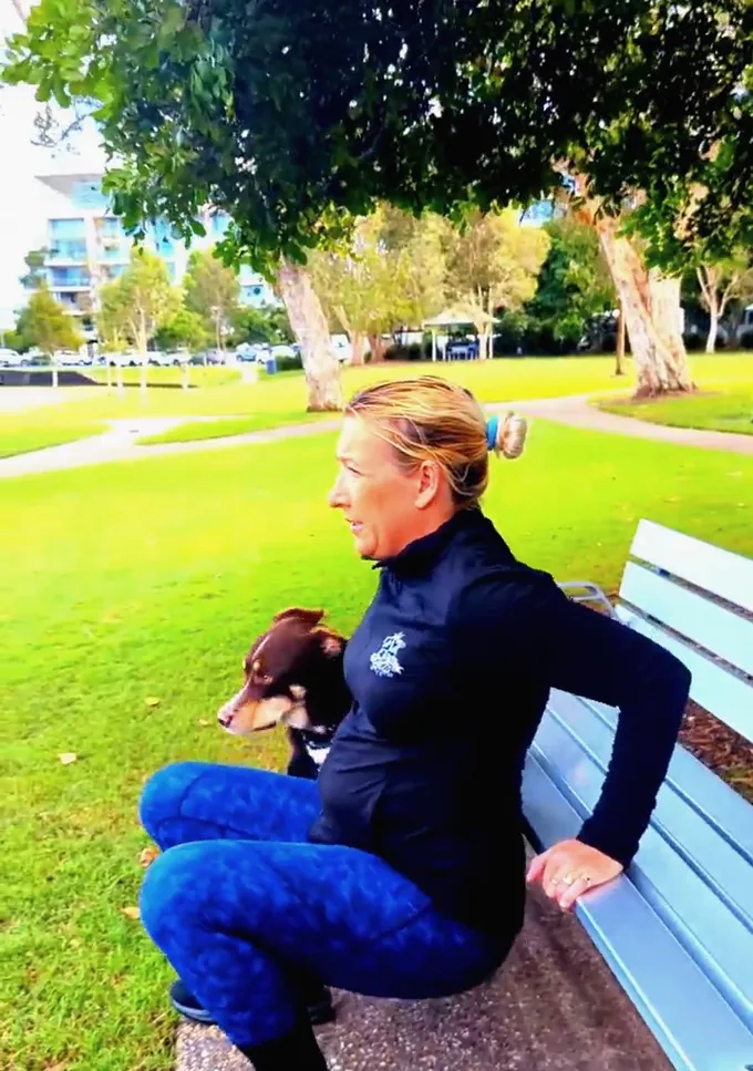 A woman doing triceps dips on a park bench with her dog sitting beside her.