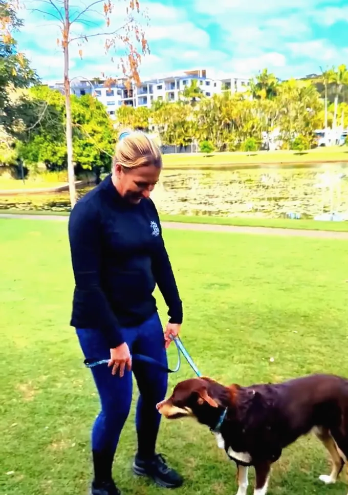 A woman doing a reverse lunge on a grassy park path with her dog watching beside her.