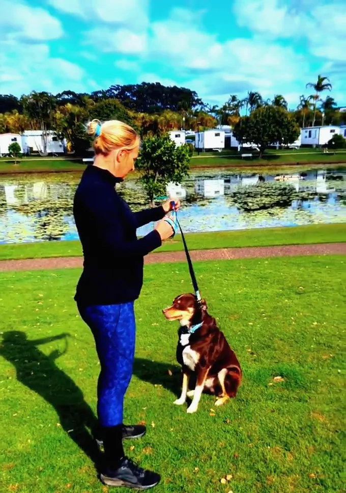 A woman doing a squat on the grass beside a lake while her dog sits next to her on the lead.
